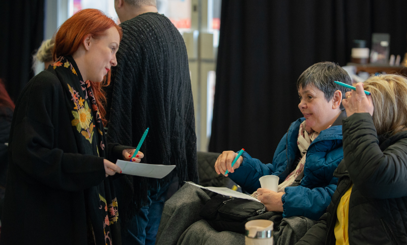 A white woman with red hair sits with a pen and notepad as she talks to a disabled woman and her support worker at an indoor event at Theatre Absolute.