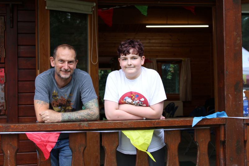 A white man and a white teenage boy stand on a the porch of a wooden cabin with bunting in front of them and smile for a photo at the camp out.