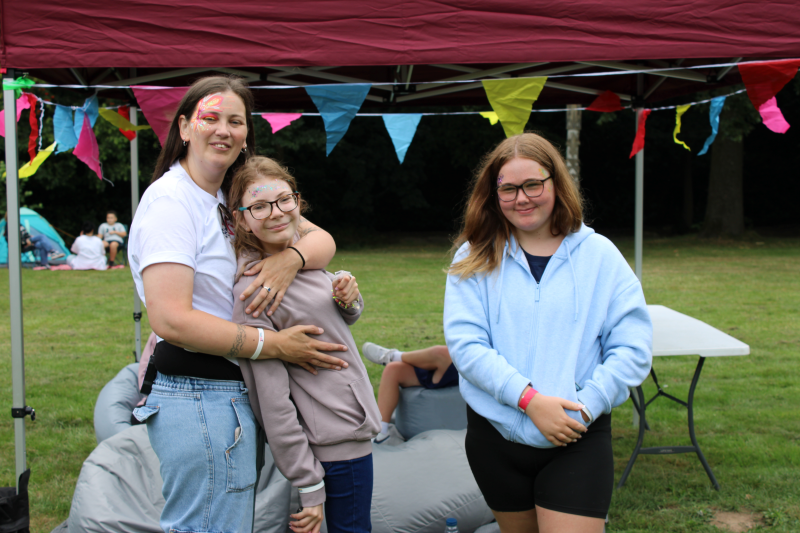 A white woman (Laura) has her arms around her daughter as they pose for a photo with another teenage girl in front of a gazebo and some colourful bunting.