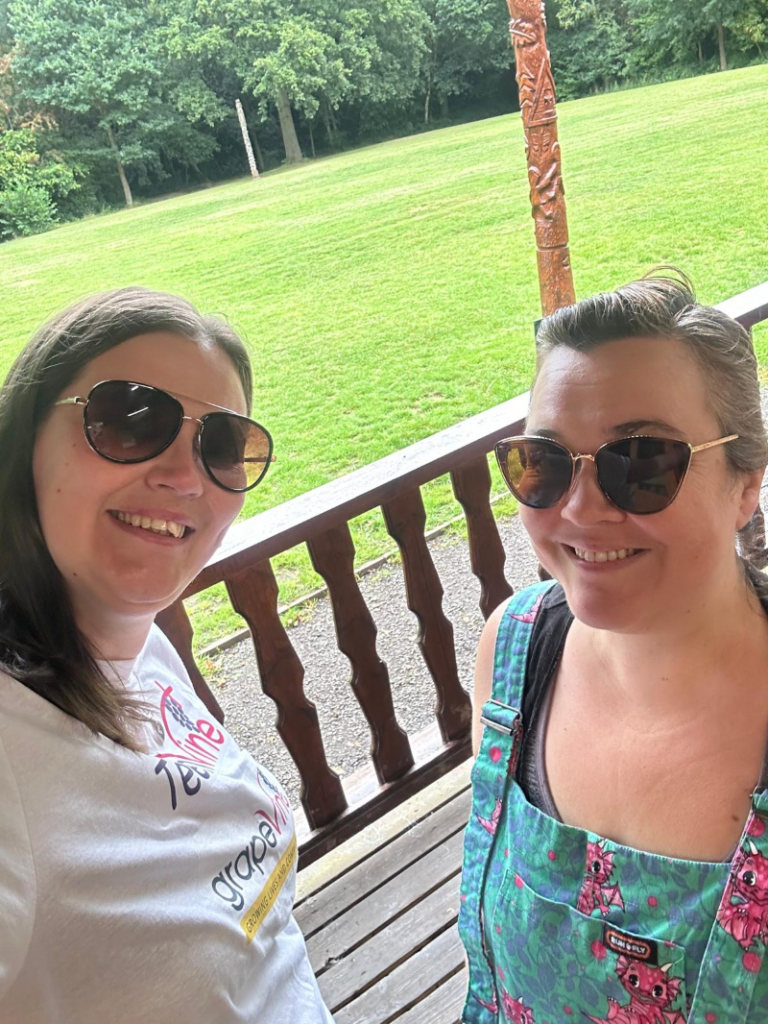 Two brunette white women, Laura and Sarah, wear sunglasses and smile for a selfie with the campsite behind them.