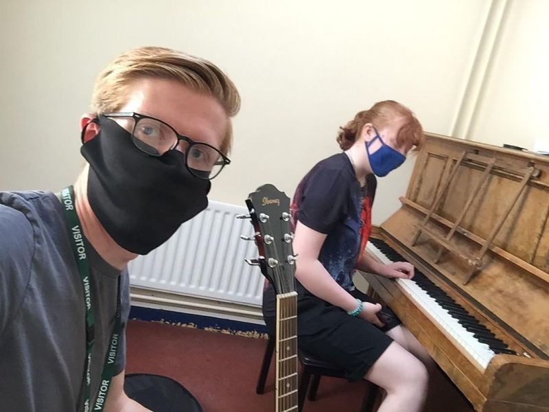 Paul from Teenvine Next Steps wears a black face mask as he sits next to a young girl wearing a blue mask and playing piano