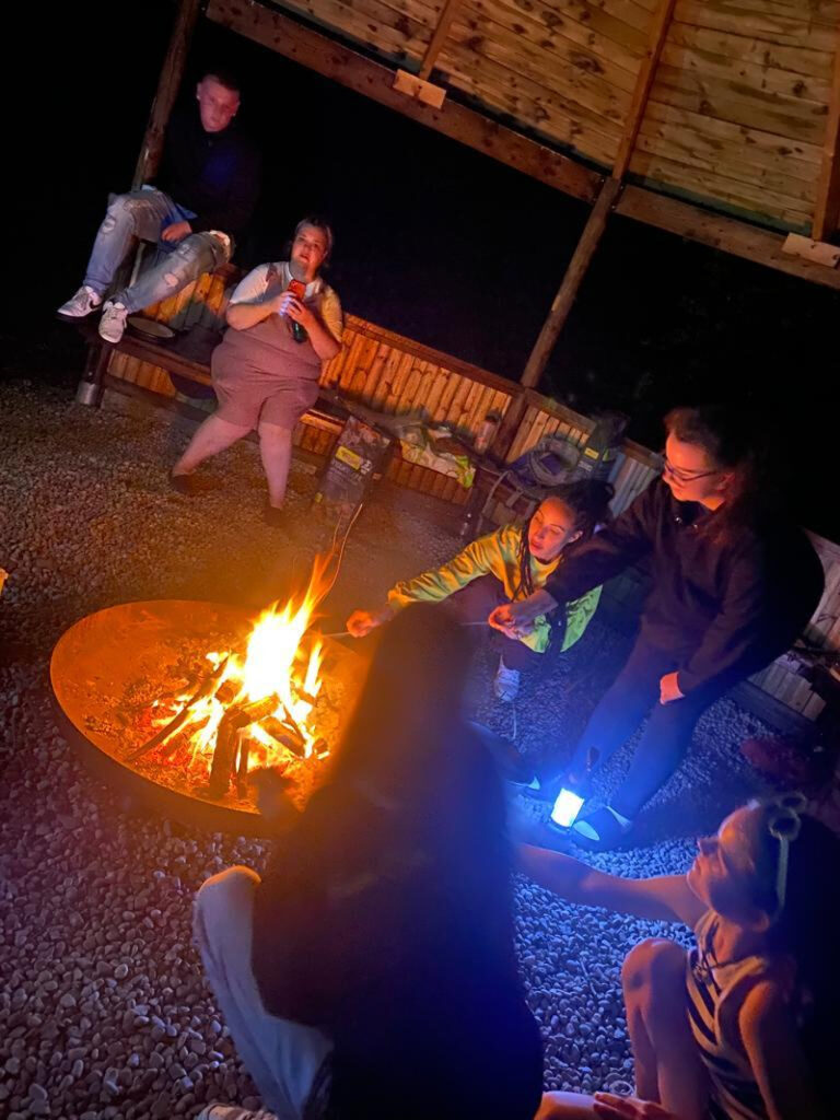 Six people, male and female, sit around a campfire under a wooden gazebo at night toasting marshmallows together.