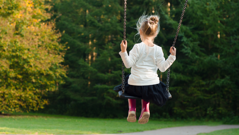 A young white girl wears a white top and pink wellies with her blonde hair as she sits on a swing in a park.