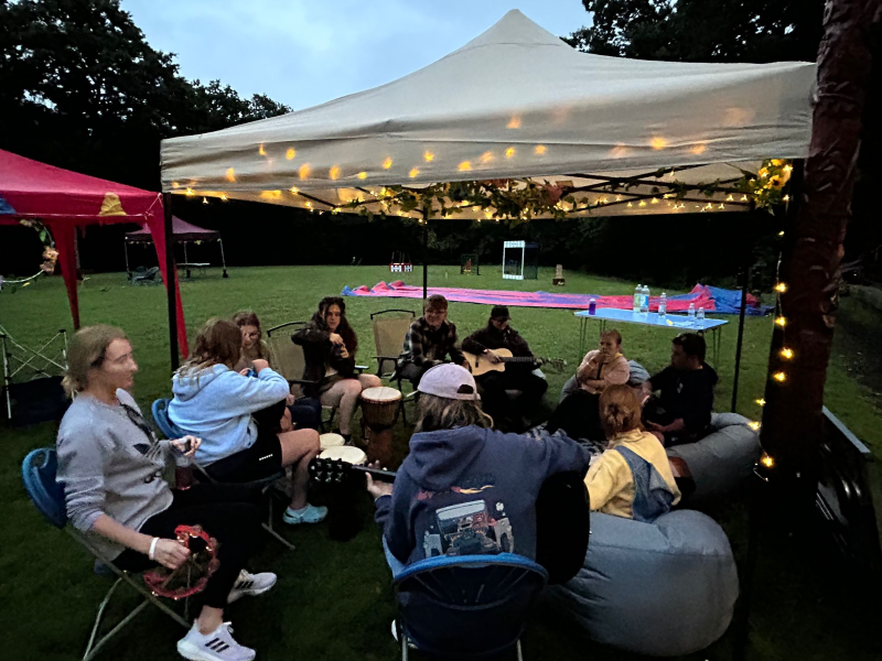 An evening at a summer festival shows young people and adults gathered beneath a white pagoda with fairy lights.