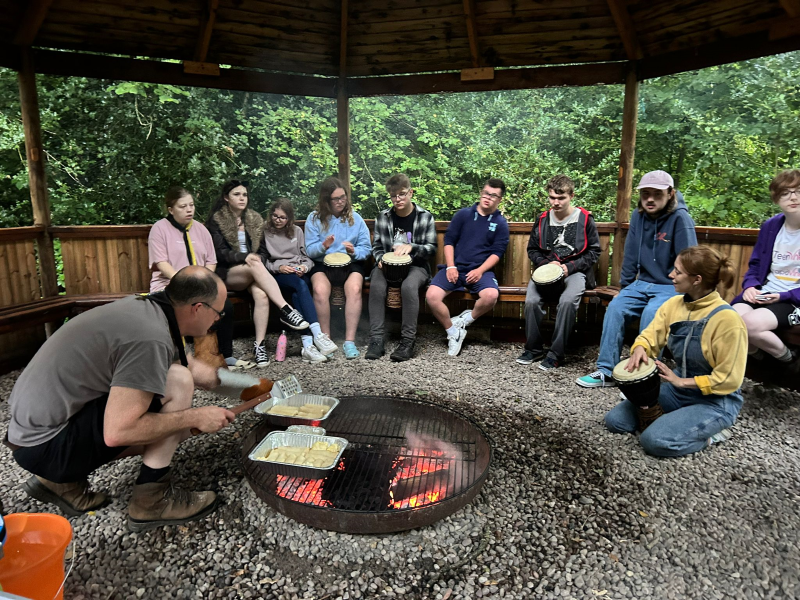 Several young people sit beneath a wooden structure around a campfire while a white man cooks sausages in two trays on the open fire.