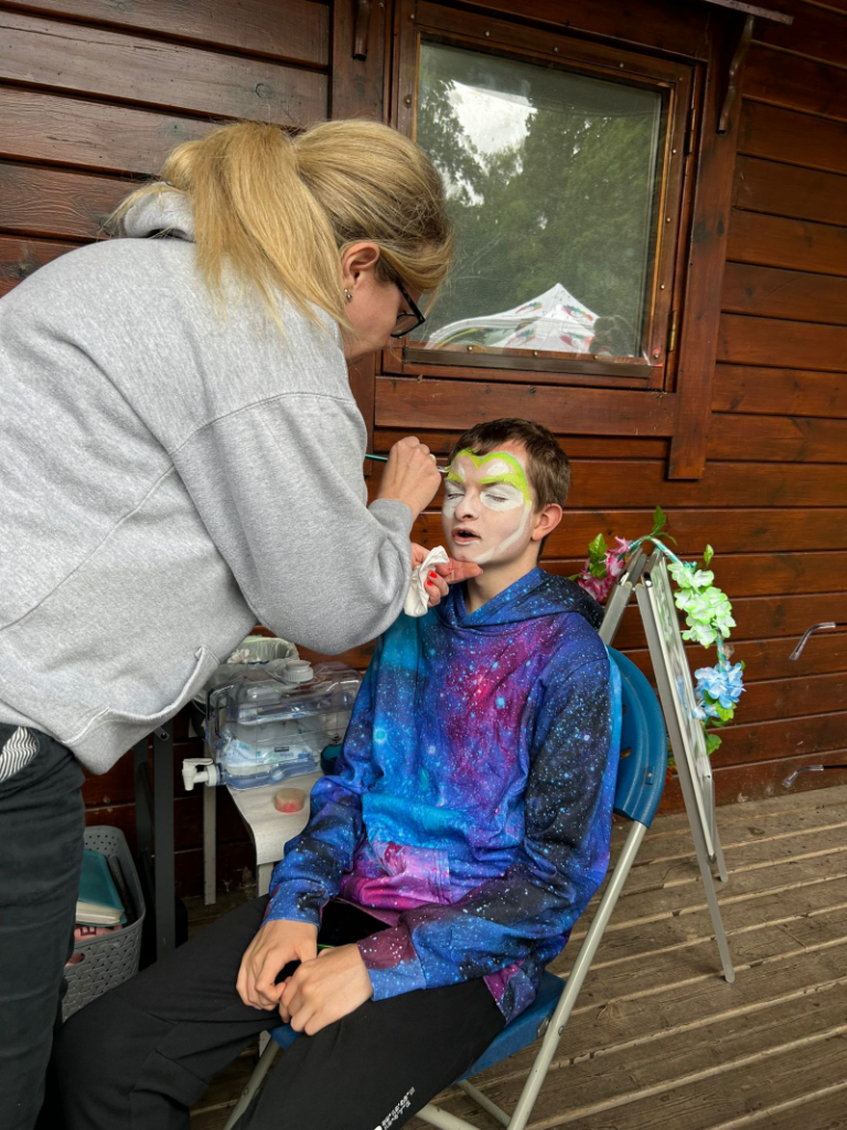 A white teenage boy wearing a blue galaxy hoodie has his face painted for the first time by a white woman with blonde hair.