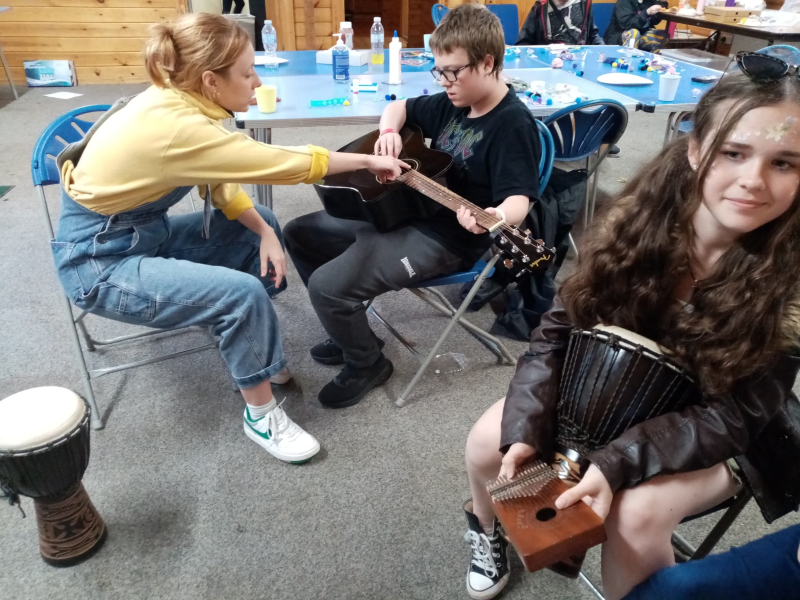 A white woman in denim dungarees shows a young white boy how to play guitar while a teenage girl looks on. They are inside the cabin.