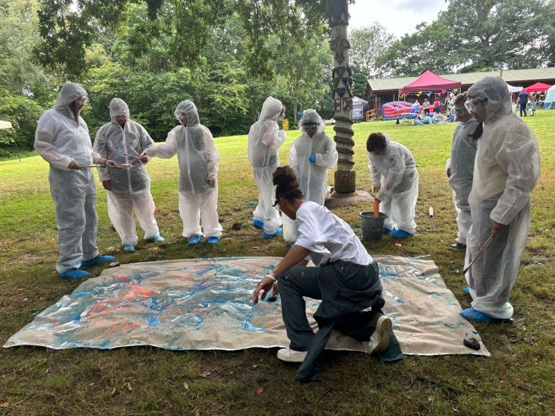 Young people in white boiler suits and goggles stand around a blank tarpaulin splashing paint on it with Naomi on a field surrounded by trees.