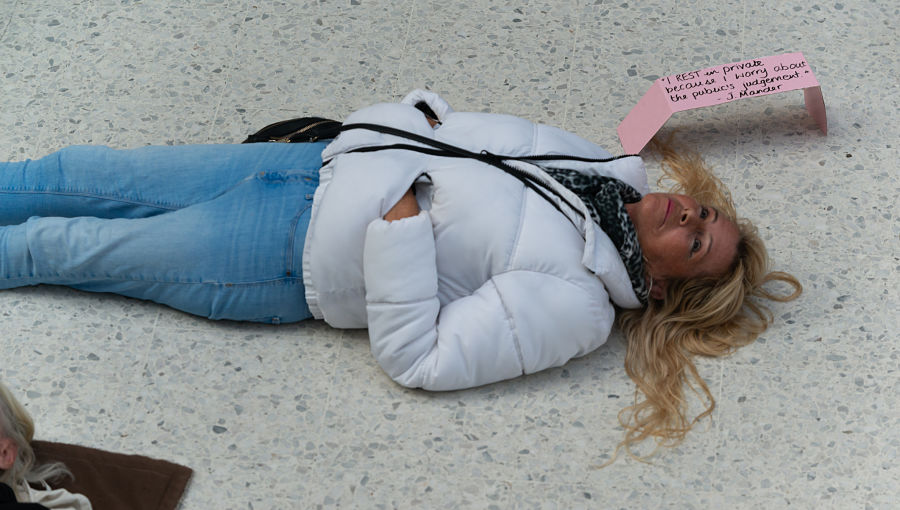 A woman in a white coat lies on the floor in a public space