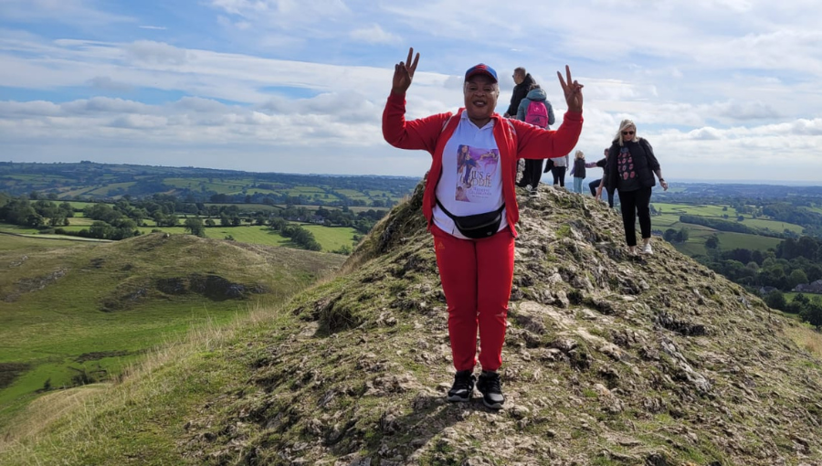 A black woman wearing a red tracksuit, bumbag and a big smile makes two peace signs with her hands on top of a hill in the Peak District on a sunny day with other walkers behind her. She is Dorothy from Waka Waka Fitness.