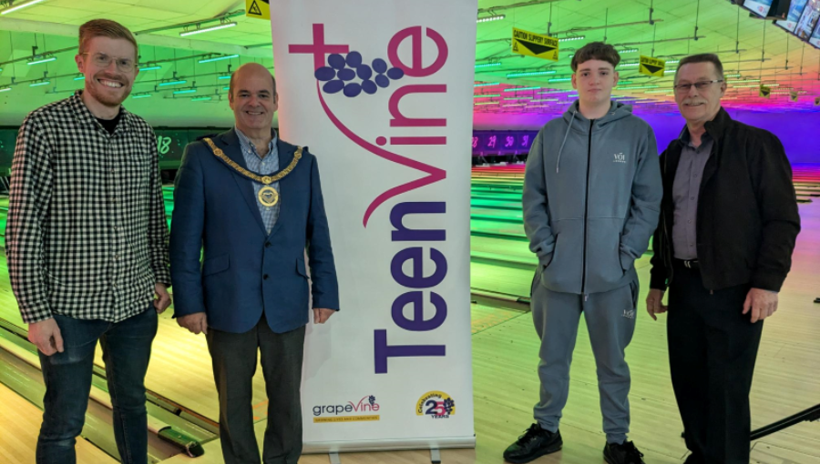 Four white males pose for a photo in a colourfully lit bowling alley in front of the Teenvine Plus promotional pull up banner. Three are adults and one is a teenager in casual clothes.