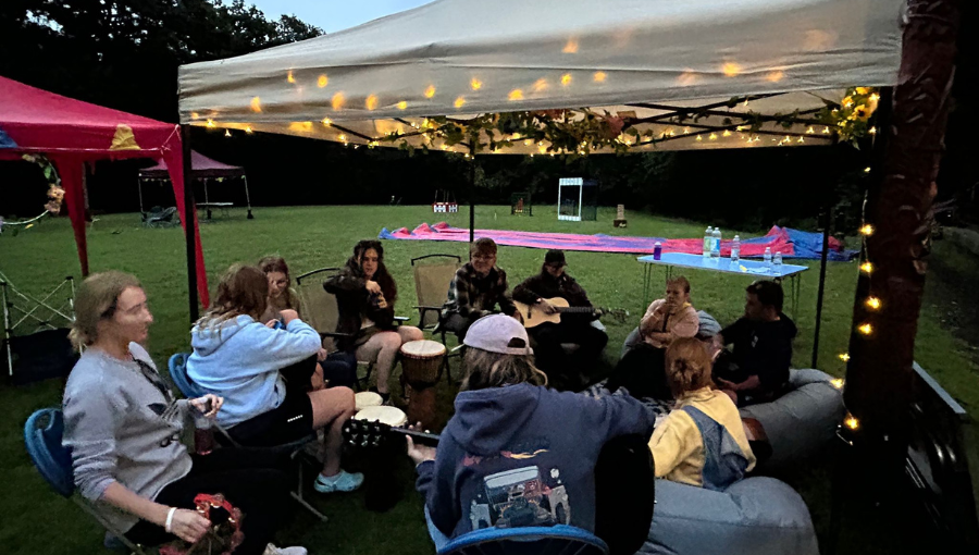 An evening at a summer festival shows young people and adults gathered beneath a white pagoda with fairy lights.