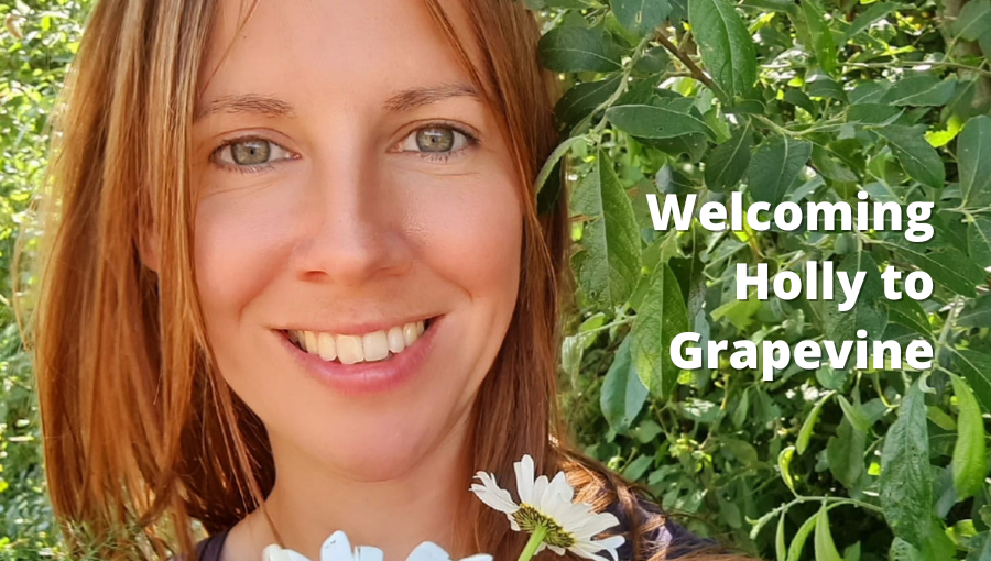 A white woman with dark blonde hair and blue eyes smiles as she holds a bunch of flowers in front of some trees in the sunshine. She is Holly Thurgood from Grapevine.