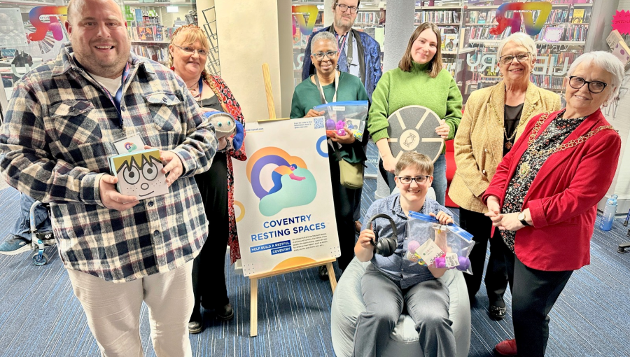 A group gathers inside the library for a publicity photo holding various sensory items available to loan and smiling for the camera.