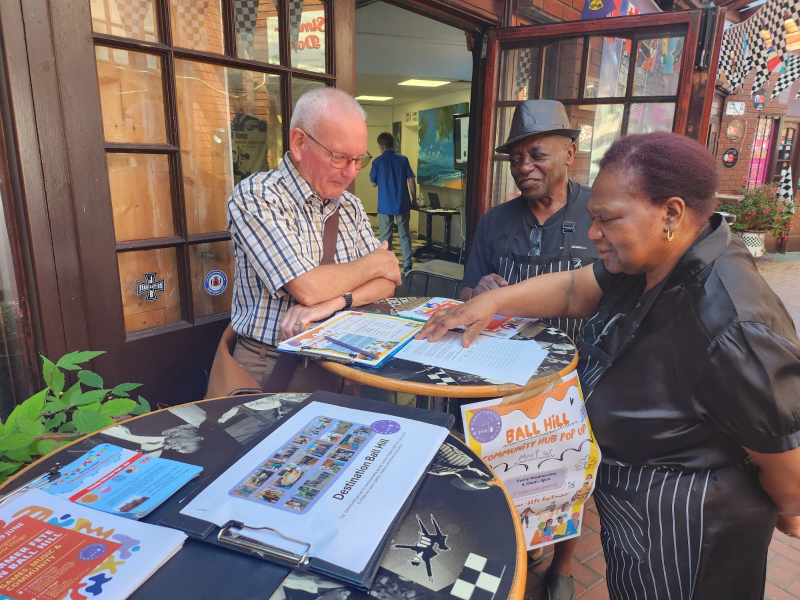 An older white man talks to an older black couple across a table containing leaflets at Ball Hill community hub which takes place every Wednesday.