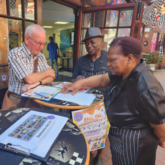 Three people - a white older man and black older couple (male and female) - stand around a round table covered in leaflets at a community event in Ball Hill, Coventry discussing what is on the table.