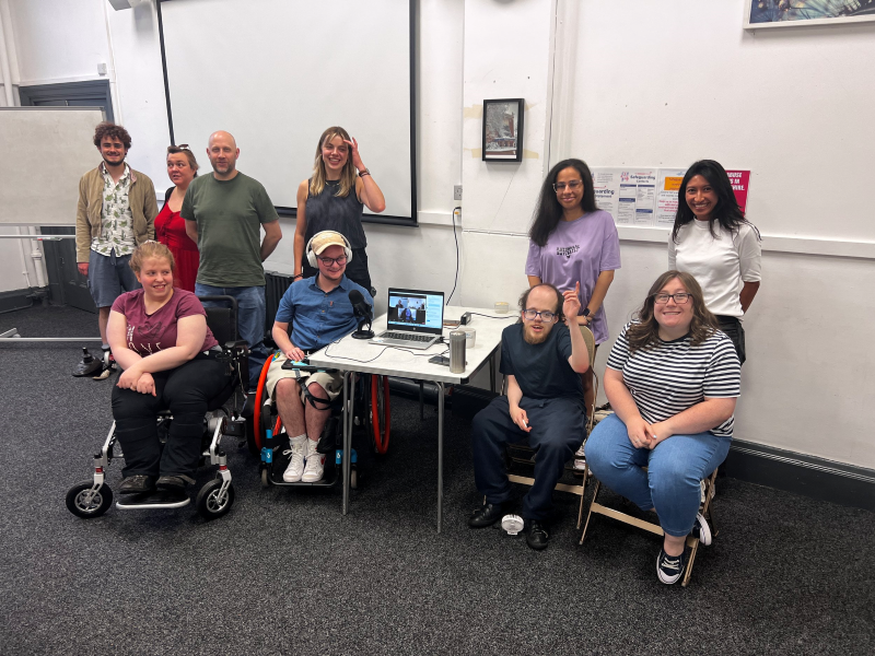 A group of people pose for a photo during a meeting. They are a mix of disabled and non-disabled people, two using wheelchairs. At the centre of the group is an open laptop.