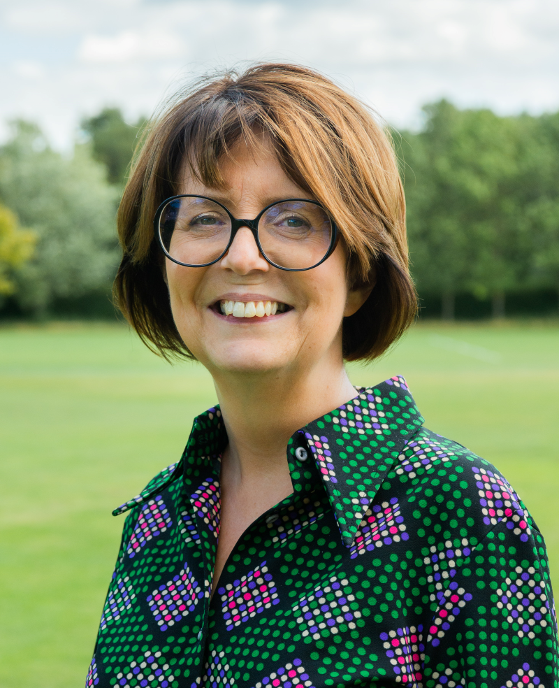 Headshot outdoors on fields at the University of Warwick of a white woman with bobbed brown hair and glasses wearing a green patterned dress and smiling. She is Clare Wightman.