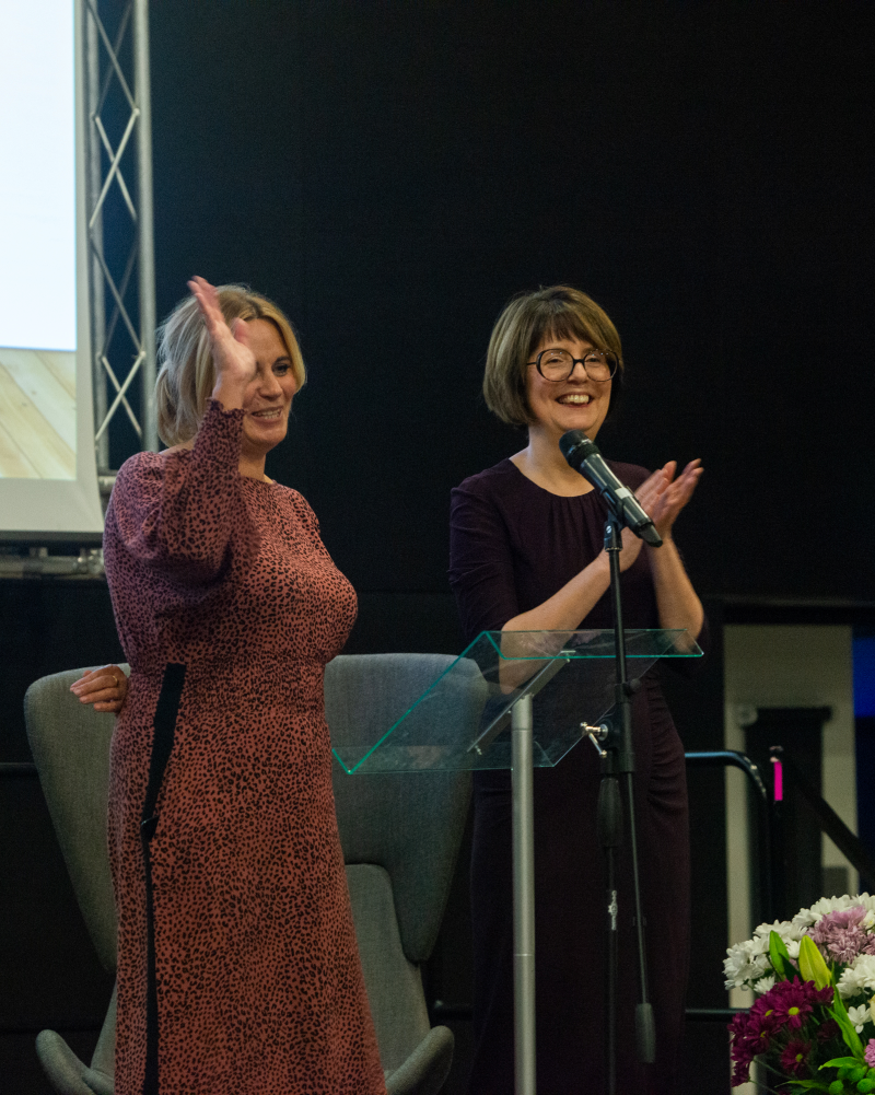 Two white women (one blonde, one brunette) smile and clap on stage at Grapevine's 25th anniversary celebration in 2019. They are Mel and Clare, Grapevine deputy CEO and CEO.