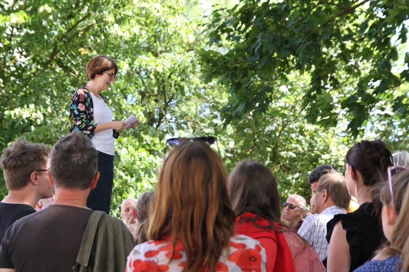 A crowd outdoors among the trees is seen listening to a white woman with bobbed brown hair and glasses (Clare Wightman) speaking at an event in Coventry city centre.