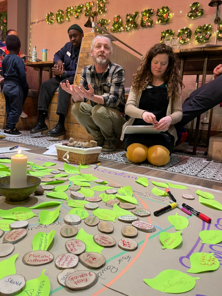 A white man with a grey beard and a white woman with brown curly hair kneel on the floor at an event and talk through the green post-its that lay on the floor next to a candle. This is a Collaboration Station event.