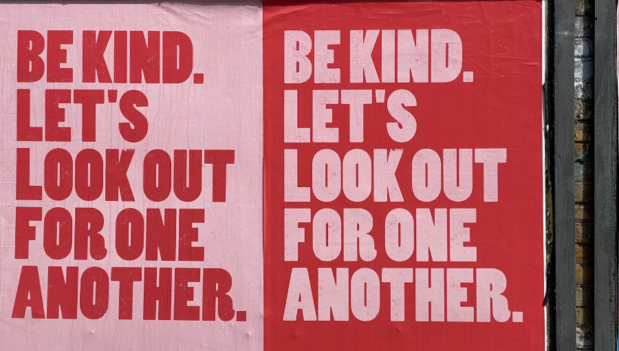 Red and pink posters pasted to an exterior wall read: Be Kind. Let's look out for one another. Image credit: John Cameron.