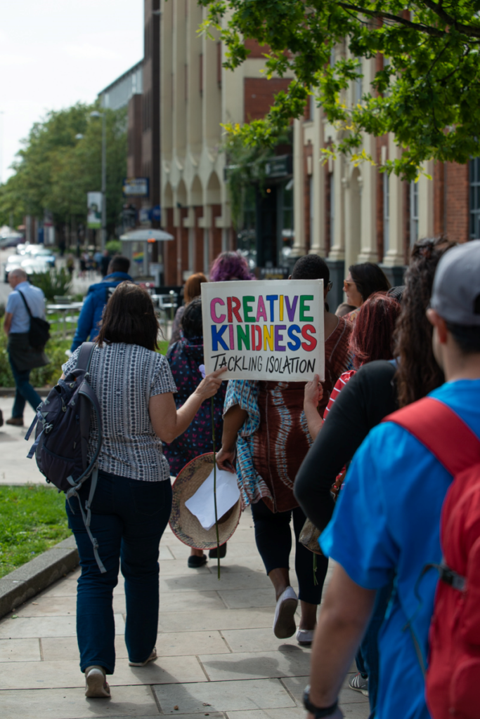 Photo taken in a city street behind a procession of people carrying placards. One says, "Creative Kindness. Tackling isolation" in bold coloured letters.