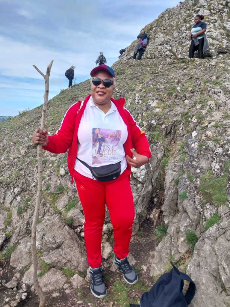 A black woman wearing a red tracksuit, bumbag and sunglasses holds a walker's wooden stick as she poses for a photo on the climb up a hill in the Peak District. She is Dorothy from Waka Waka Fitness.