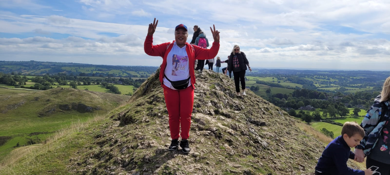 A black woman wearing a red tracksuit, bumbag and a big smile makes two peace signs with her hands on top of a hill in the Peak District on a sunny day with other walkers behind her. She is Dorothy from Waka Waka Fitness.