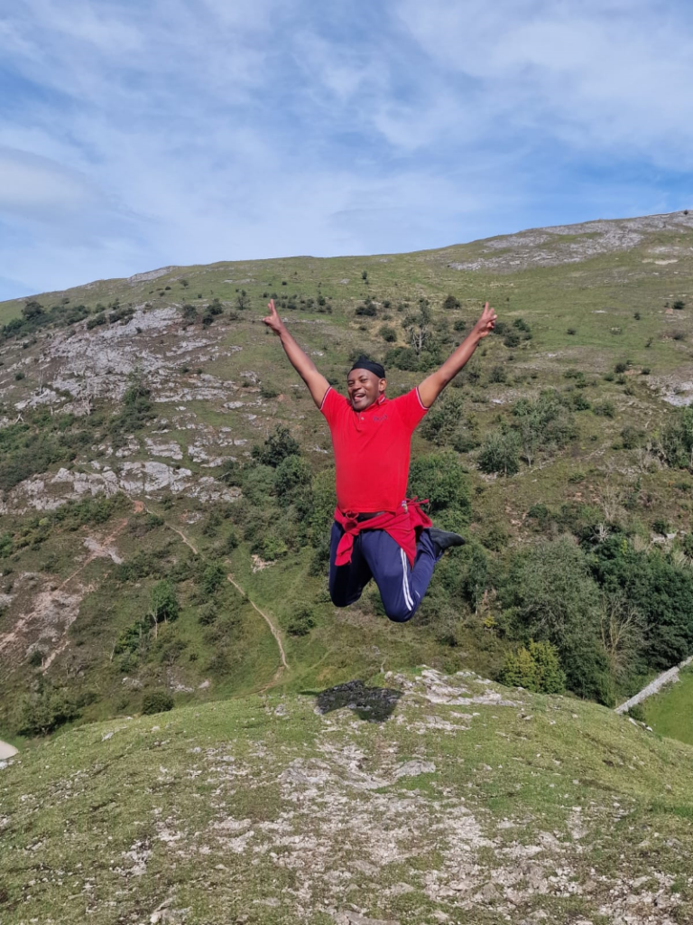 Grapevine community organiser Edwin, a black man in a red t-shirt, jumps for joy on the hillside.