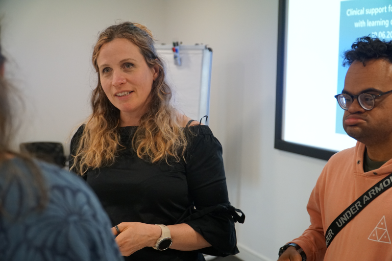 A white woman with long blonde wavy hair stands next to a mixed race man who has Down's syndrome as they talk to an NHS staff member in a training room.