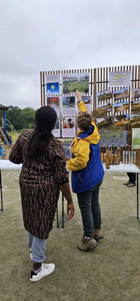 Two women, one white and one black, are turned away from the camera as they examine an information board with development plans and photos on it at a local Coventry park.