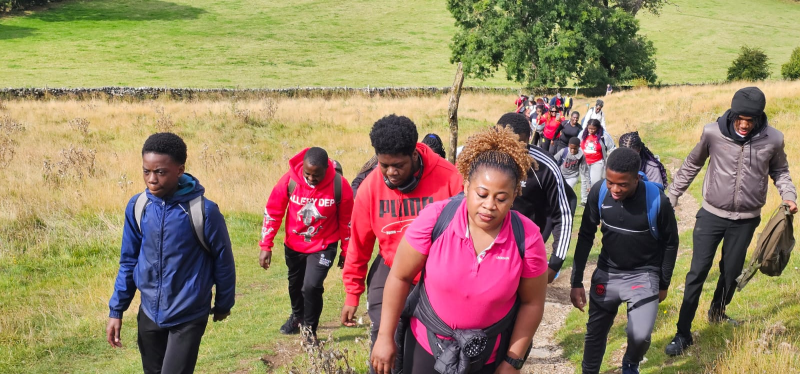 A mixed group of men and women, all black, wearing comfortable clothing and rucksacks to climb a hill in the Peak District together.