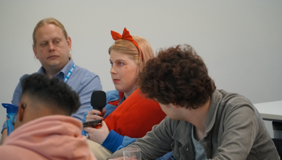 A young blonde woman wears a red bow in her hair and holds a microphone as she addresses doctors and nurses on a training course.