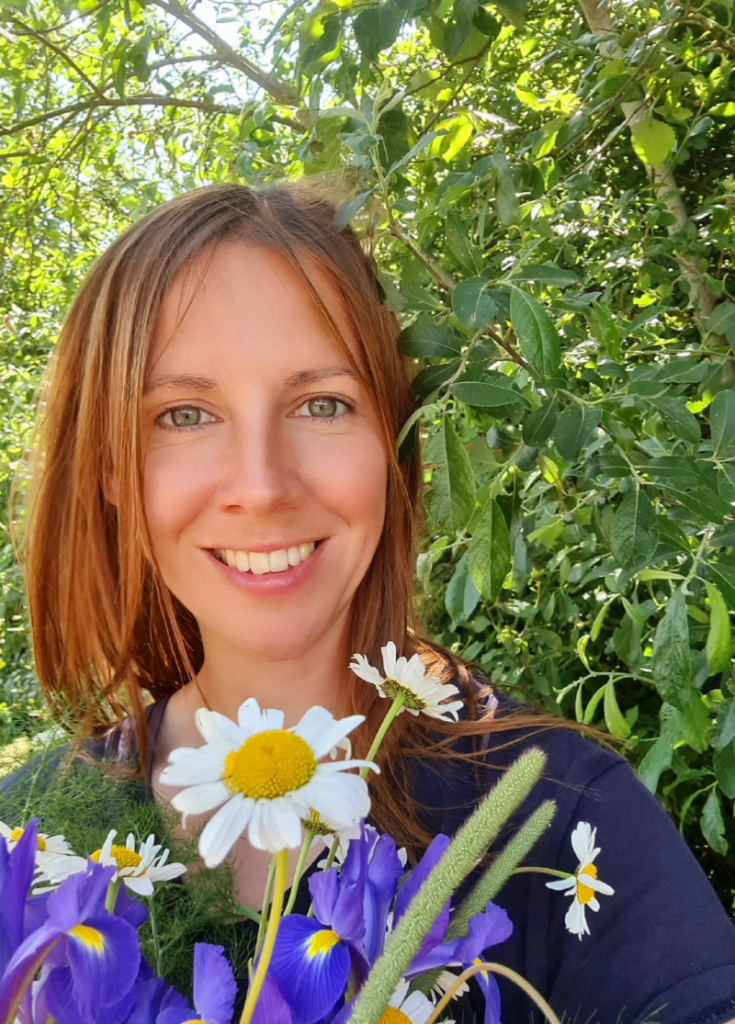 A white woman with dark blonde hair and blue eyes smiles as she holds a bunch of flowers in front of some trees in the sunshine. She is Holly Thurgood from Grapevine.