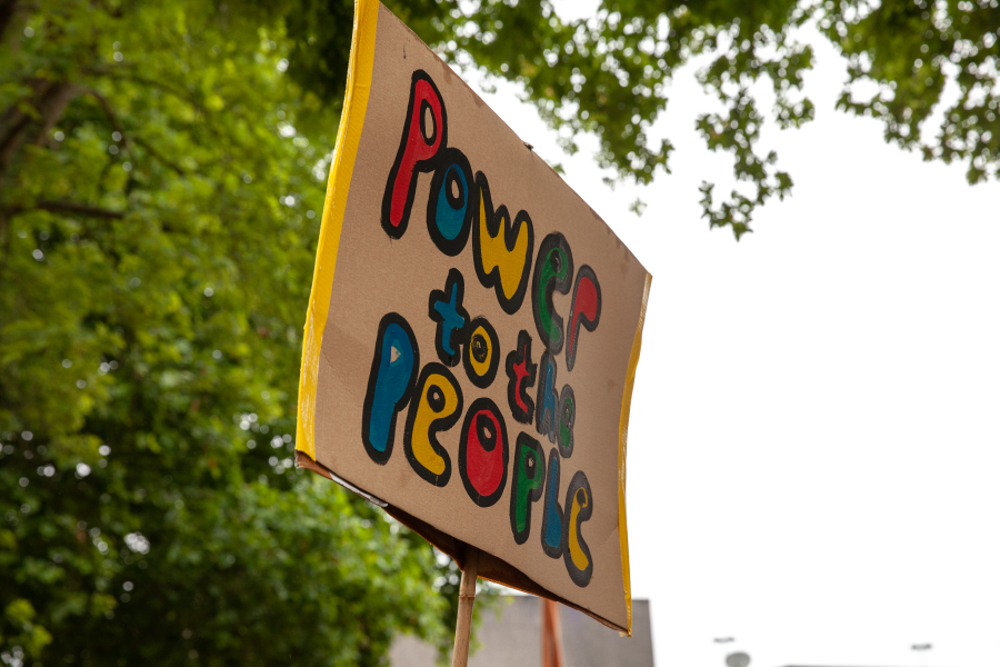 A colourful handmade placard on cardboard reading 'Power to the People' is held high in front of some trees and a grey sky.