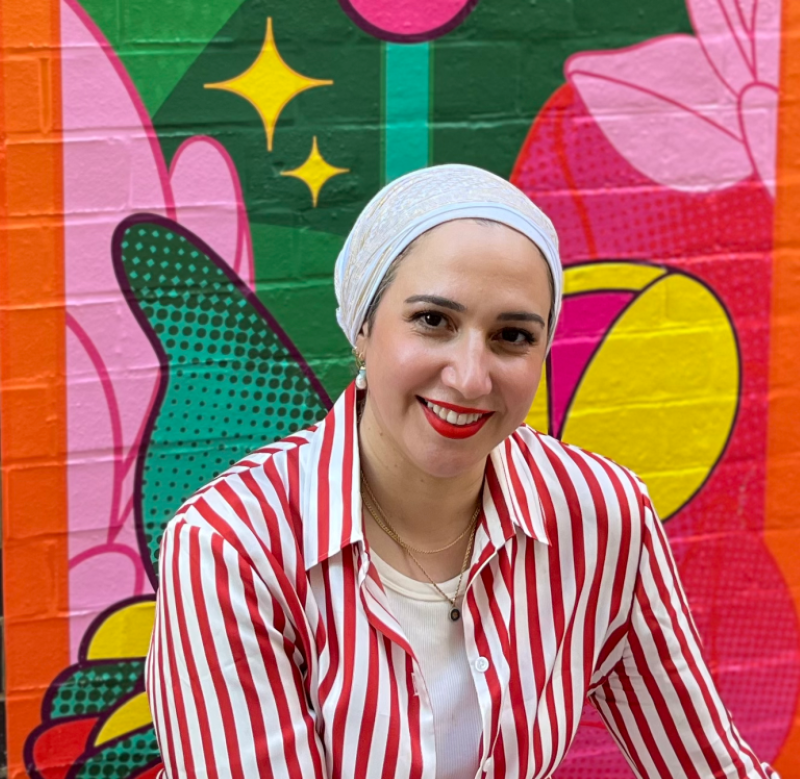 Reem, a Middle Eastern woman wearing a beige scarf to cover her hair and a red and white pinstripe shirt over a white t-shirt, smiles for a portrait photo in front of a brightly painted brick wall.