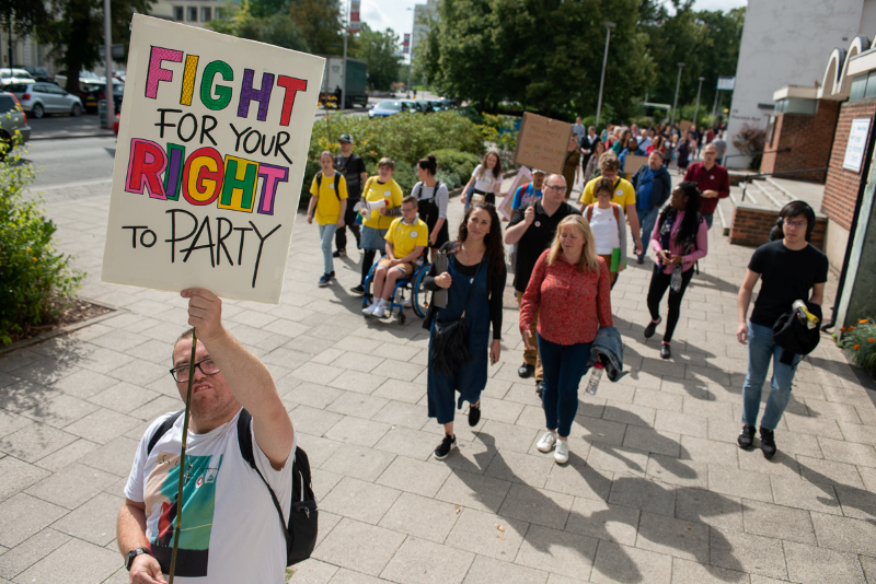 A white disabled man wears a white t-shirt on a sunny day and holds a placard aloft that reads, "Fight for your right to party" in bold coloured letters. Behind him a long line of people follow through city centre streets.