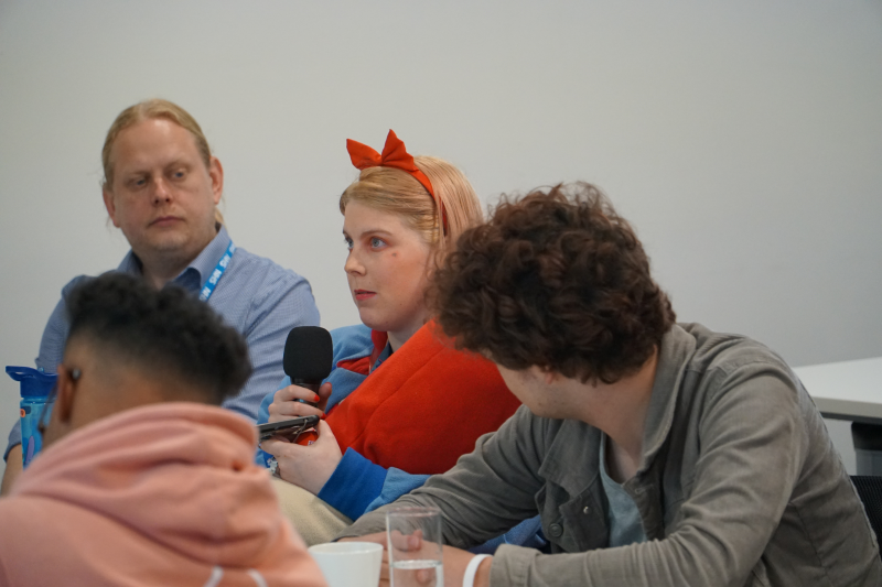 A young blonde woman wears a red bow in her hair and holds a microphone as she addresses doctors and nurses on a training course.