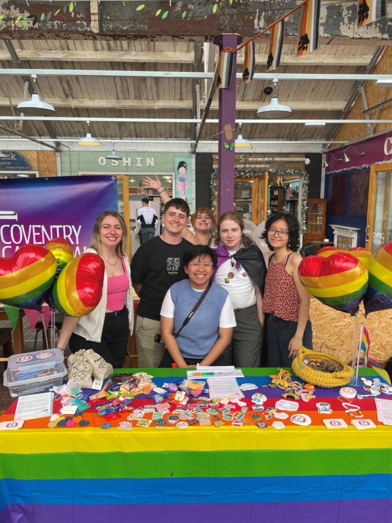 A group of six smiling people stand behind an indoor information stall that is covered in rainbows.