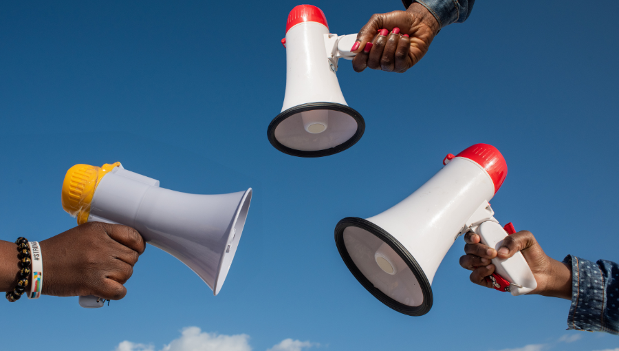 A stock photo shows three black adult hands each holding a red and white megaphone in the air against a blue sky.