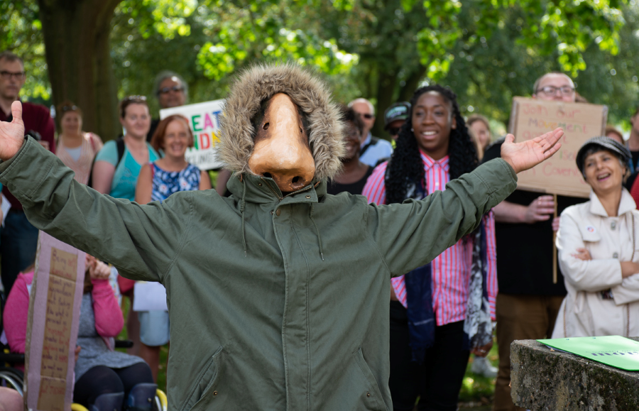 A man wearing a green parker coat with his whole face covered by a giant nose mask, spreads his arms wide as crowds behind him look on and smile.